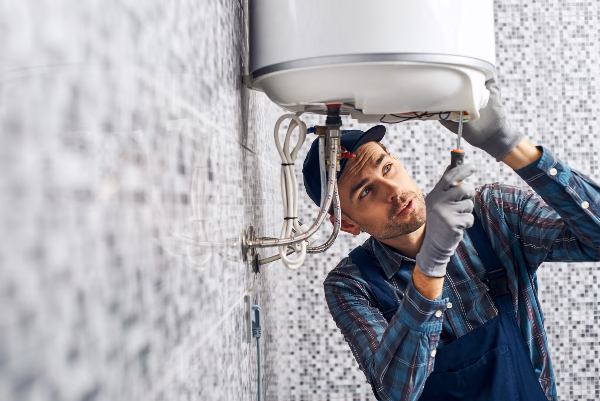 Technician wearing gloves and a cap uses a screwdriver to repair a wall-mounted water heater.