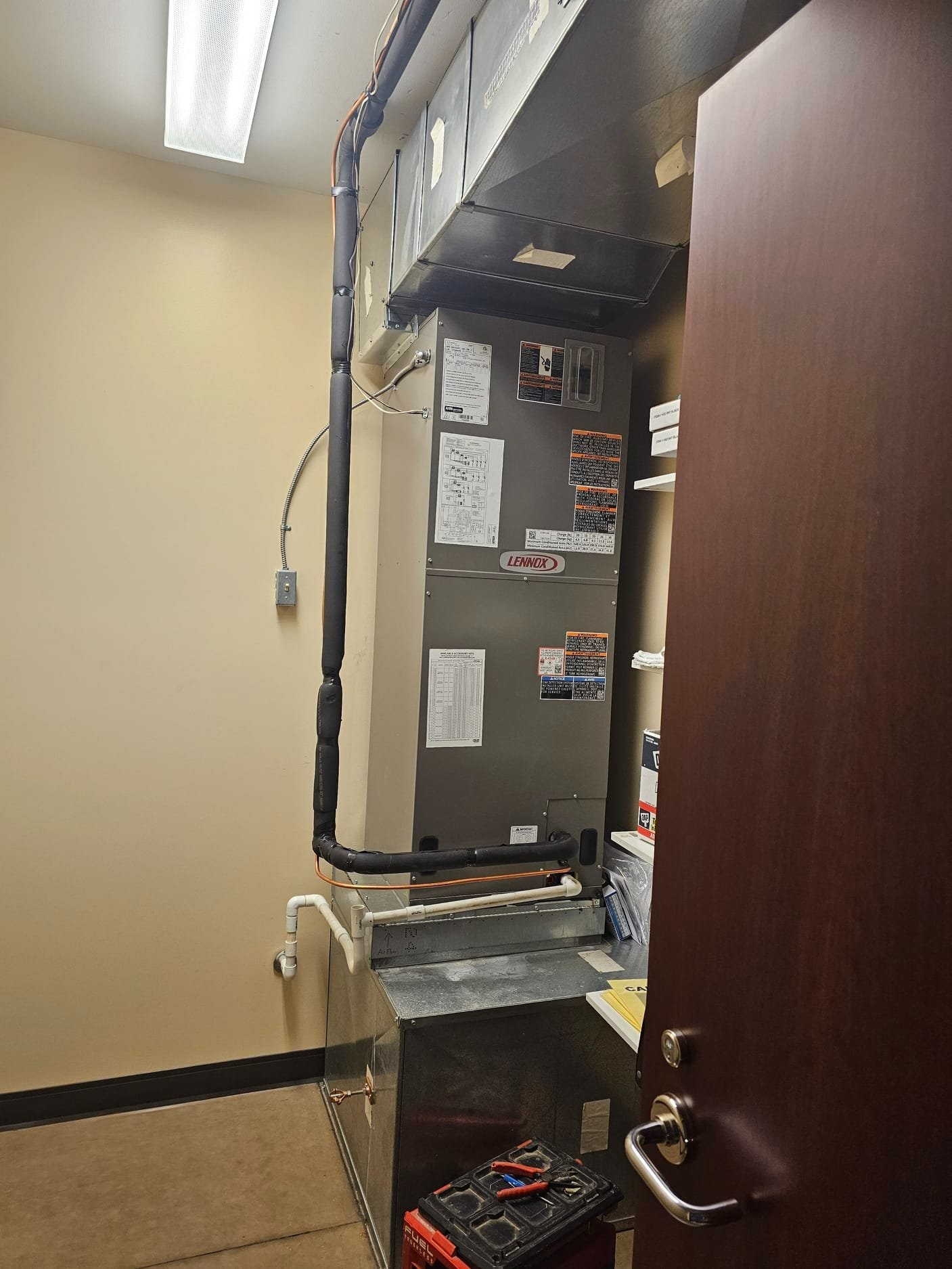 Indoor gray Lennox heating and air conditioning unit with insulated pipes, tools on a toolbox, and a partially open dark wooden door.
