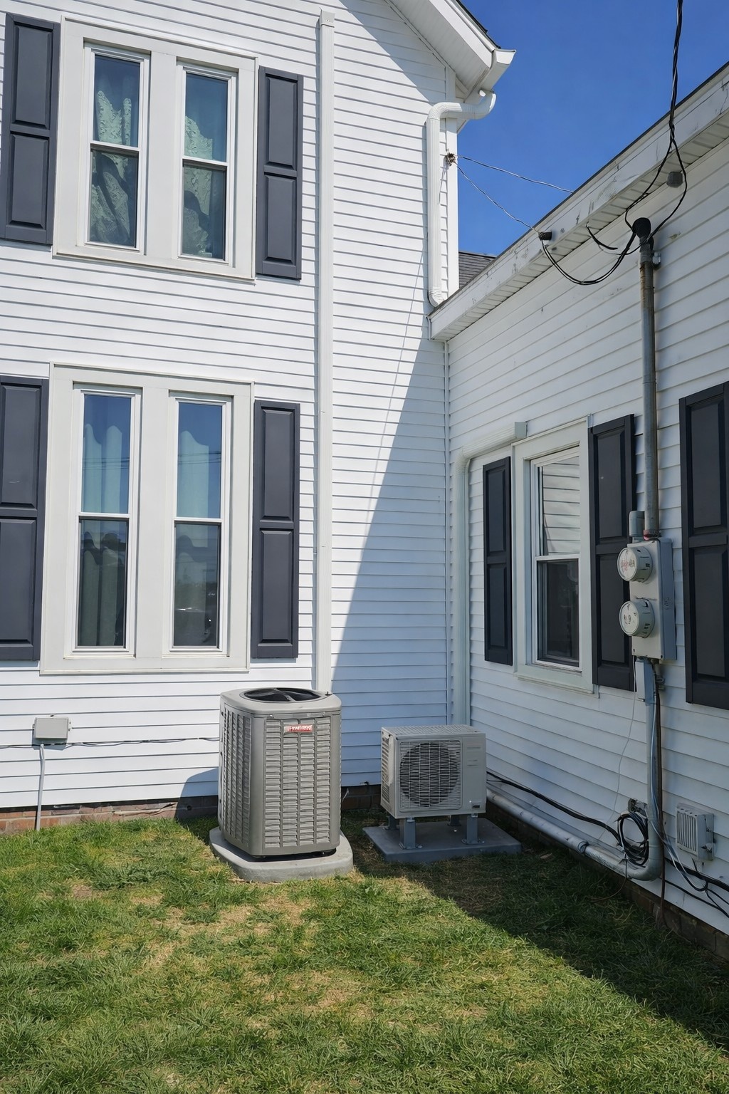Two air conditioning units installed outside a white house with black window shutters on a sunny day.