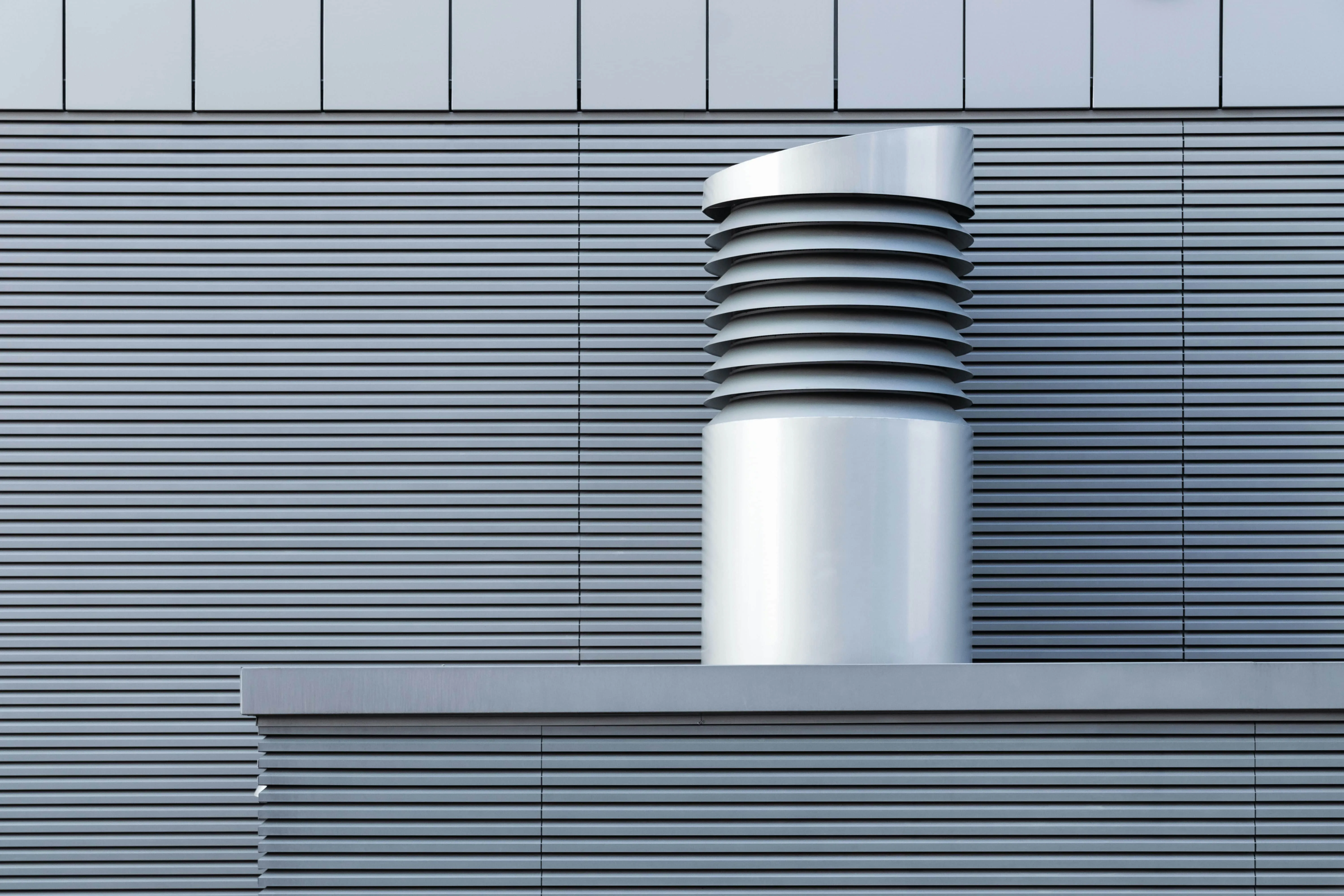 Modern architectural detail of a silver ventilation system on a ribbed gray building facade.
