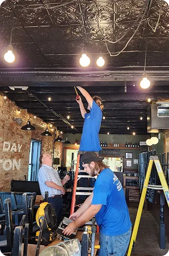 Workers performing maintenance in a restaurant with exposed brick walls and warm lighting.
