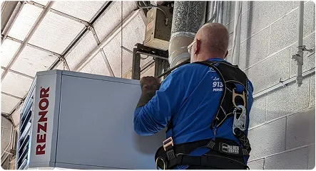 A technician adjusts a REZNOR heater while using safety harnesses in a warehouse environment.
