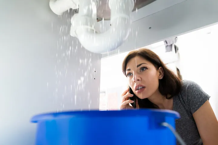 A concerned woman talks on her phone while watching water leak from a sink into a blue bucket.
