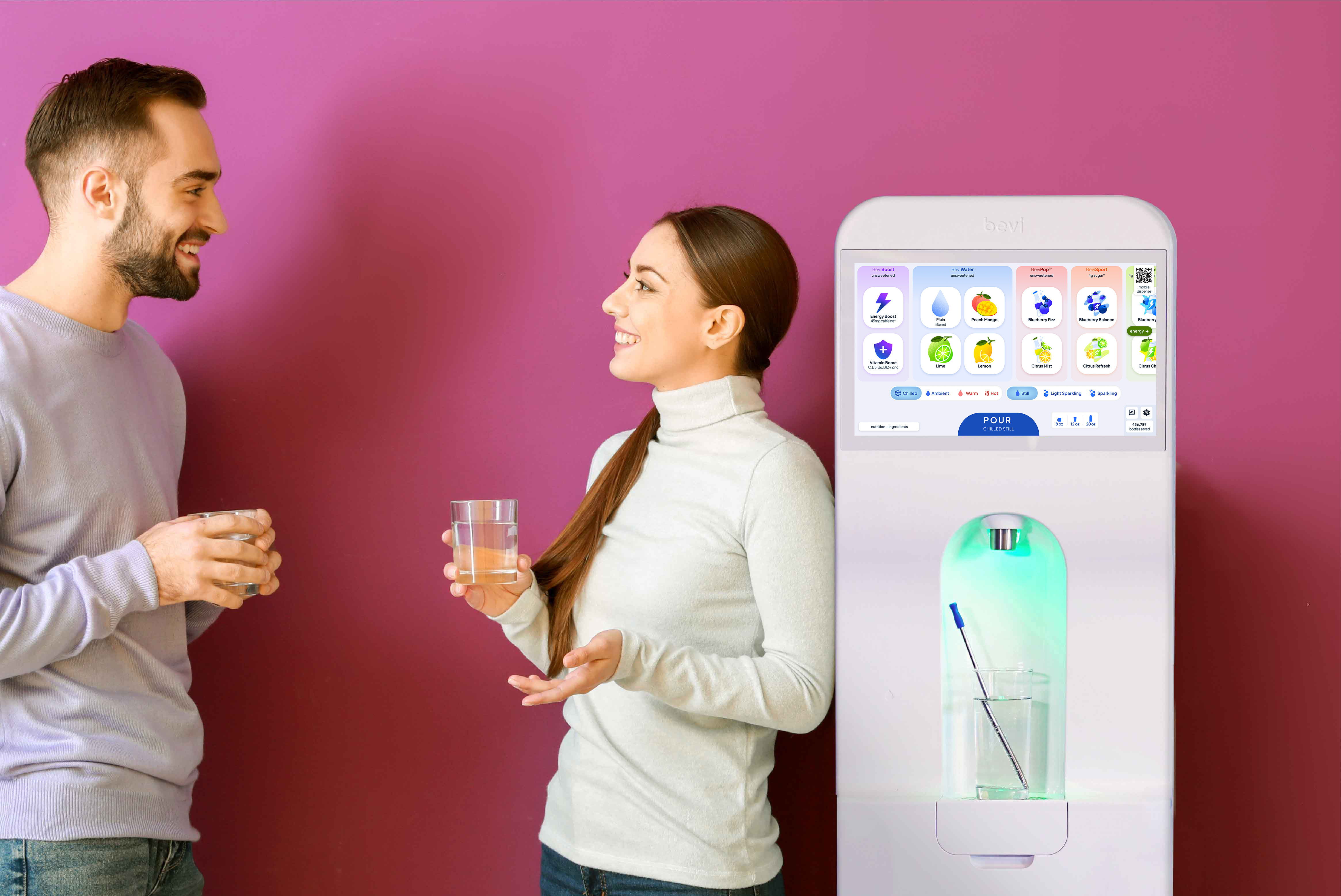 Two employees smile at each other while holding glasses of sparkling water beside a Bevi 2.0.