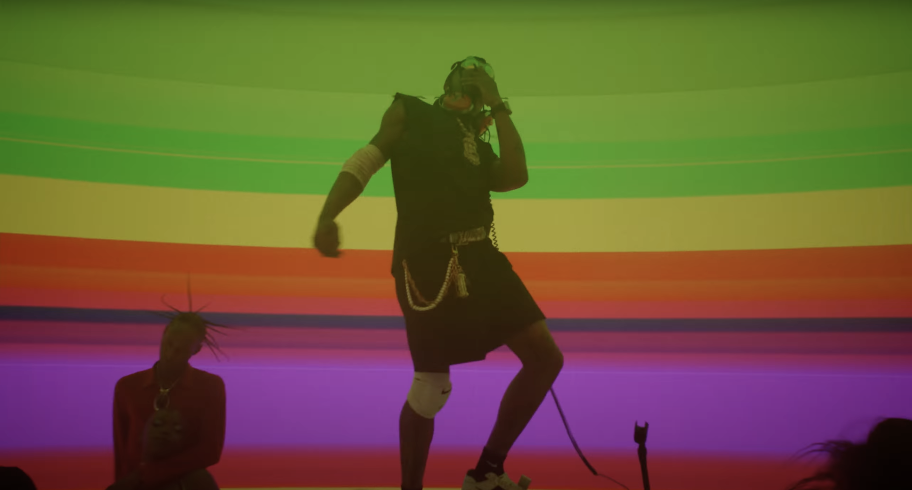 Performer on stage with colorful horizontal rainbow stripes background singing into a microphone. - Screenshot of the videoclip TRAVIS SCOTT, Modern Jam