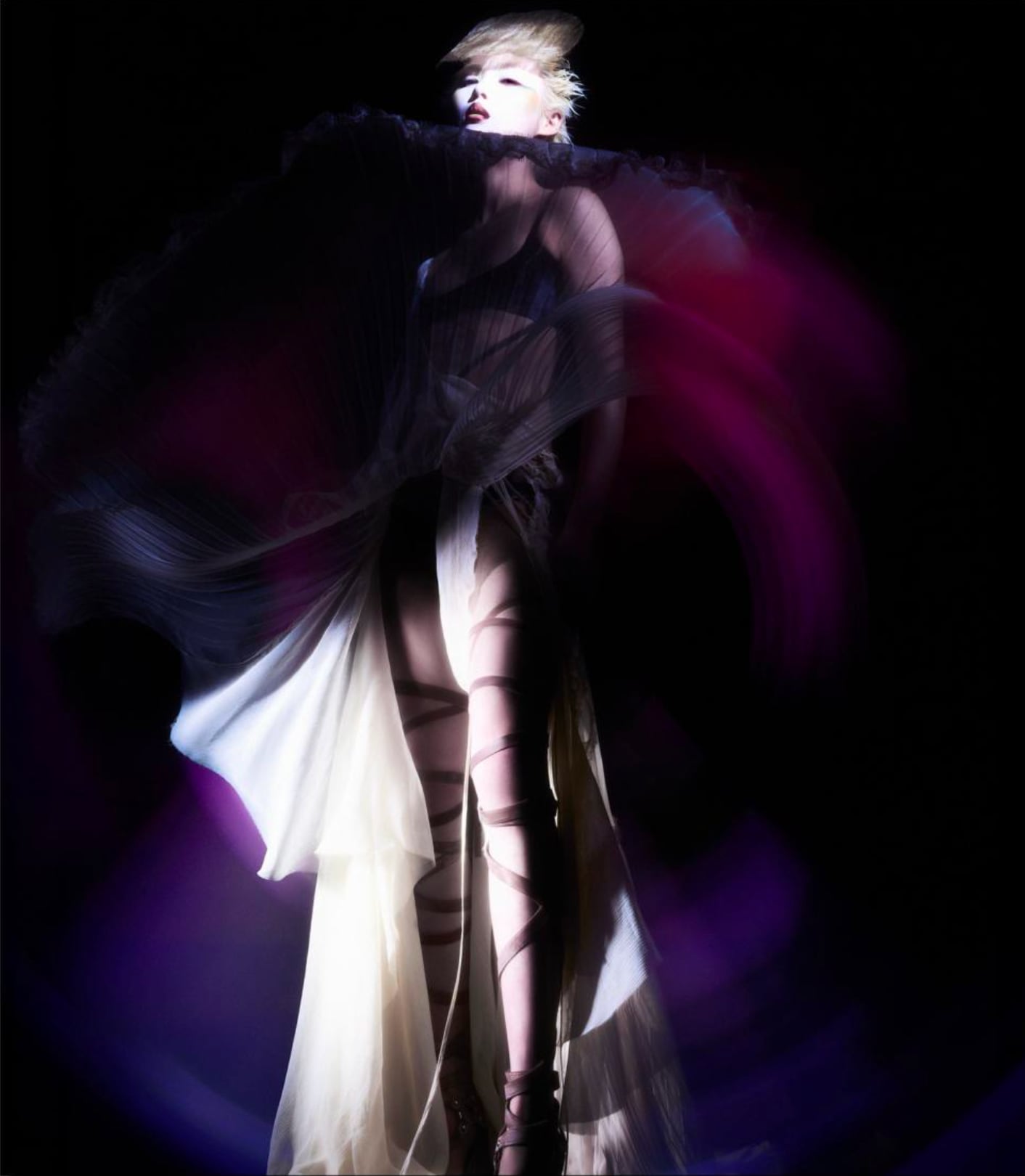 Fashion model in dramatic pose wearing a flowing black and white dress with sheer fabric and ribbons, illuminated against a dark background with purple and pink light swirls. - from the Magazine NUMÉRO Aurora 12 - Lure of the sea