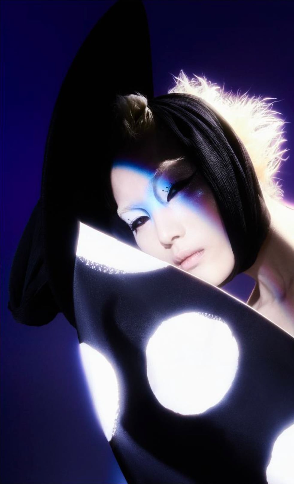 Portrait of a person with dramatic makeup and a black garment with large white dots, partially covering their face, set against a dark purple background. - from the Magazine NUMÉRO Aurora 12 - Lure of the sea
