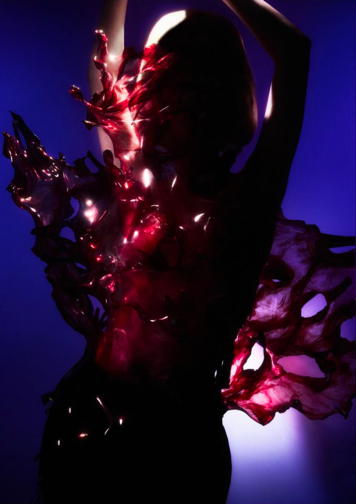 Silhouette of a person with arms raised wearing a dark translucent, textured garment glowing with red highlights against a blue background. - from the Magazine NUMÉRO Aurora 12 - Lure of the sea