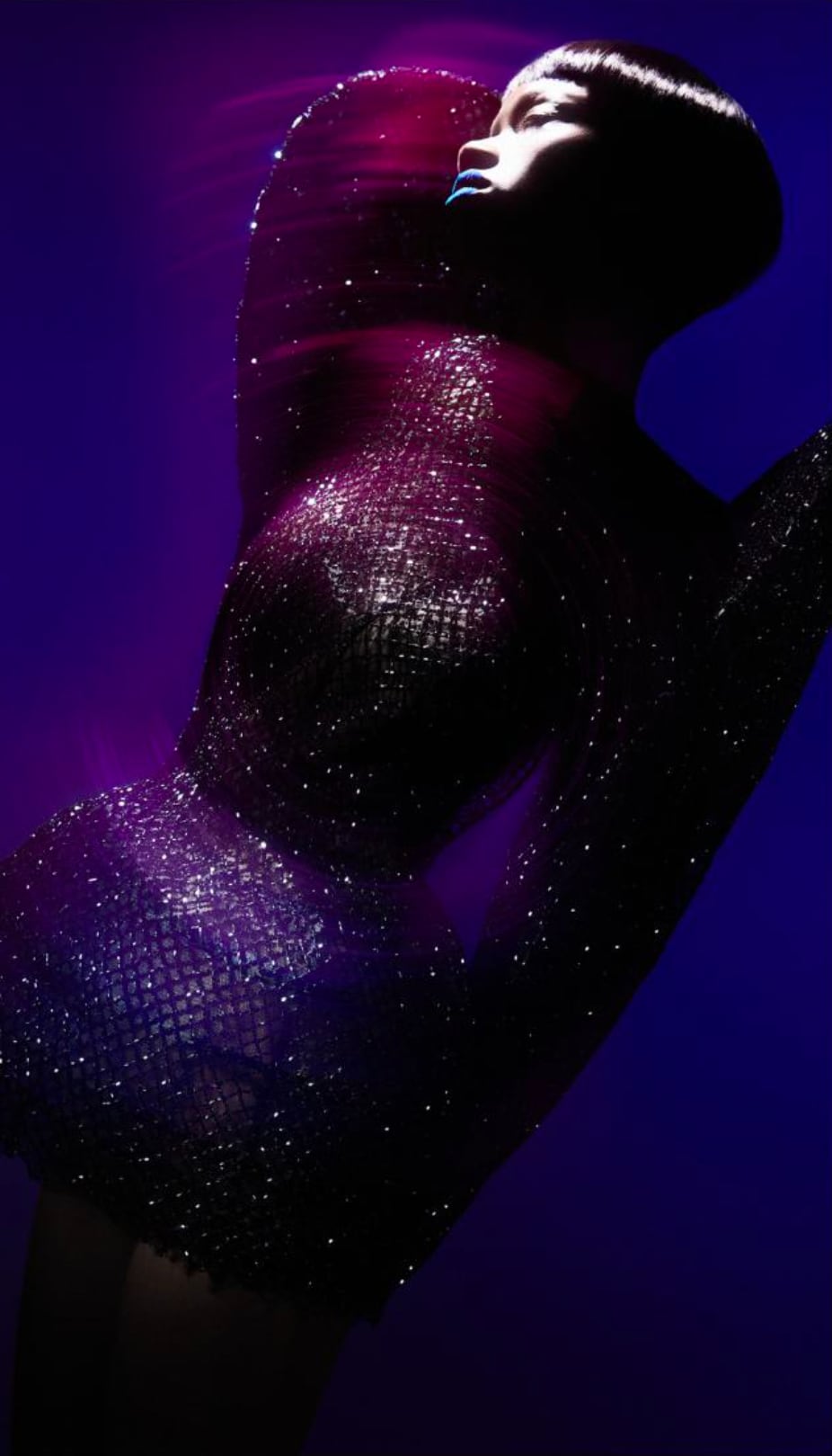 Side profile of a person with short hair wearing a glittery, dark textured outfit illuminated by purple and blue light on a dark background. - from the Magazine NUMÉRO Aurora 12 - Lure of the sea