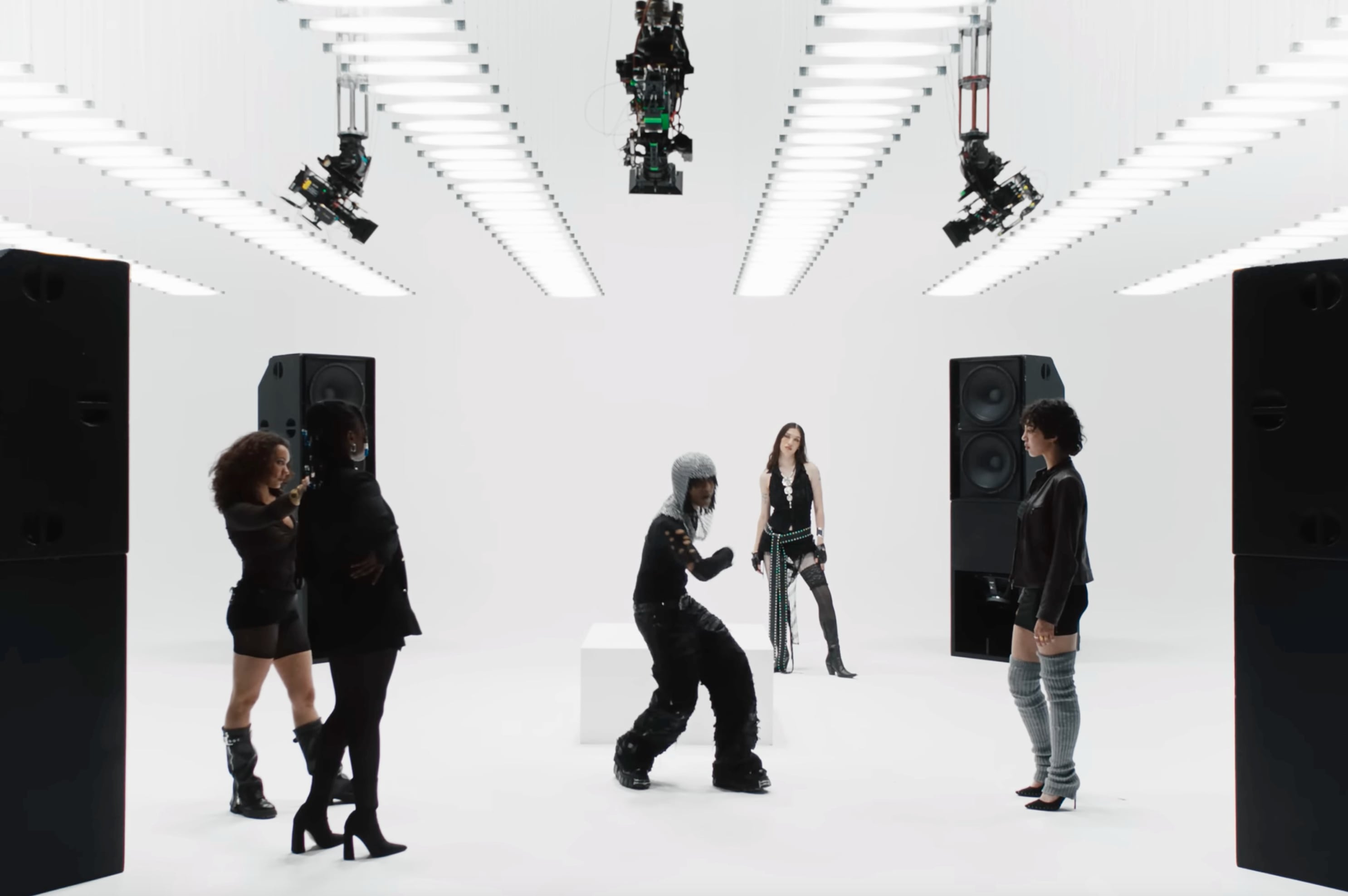 Five people dressed in black standing and dancing in a bright white studio with large speakers and overhead lights. - screenshot of the videoclip ADES THE PLANET, 24 Carats