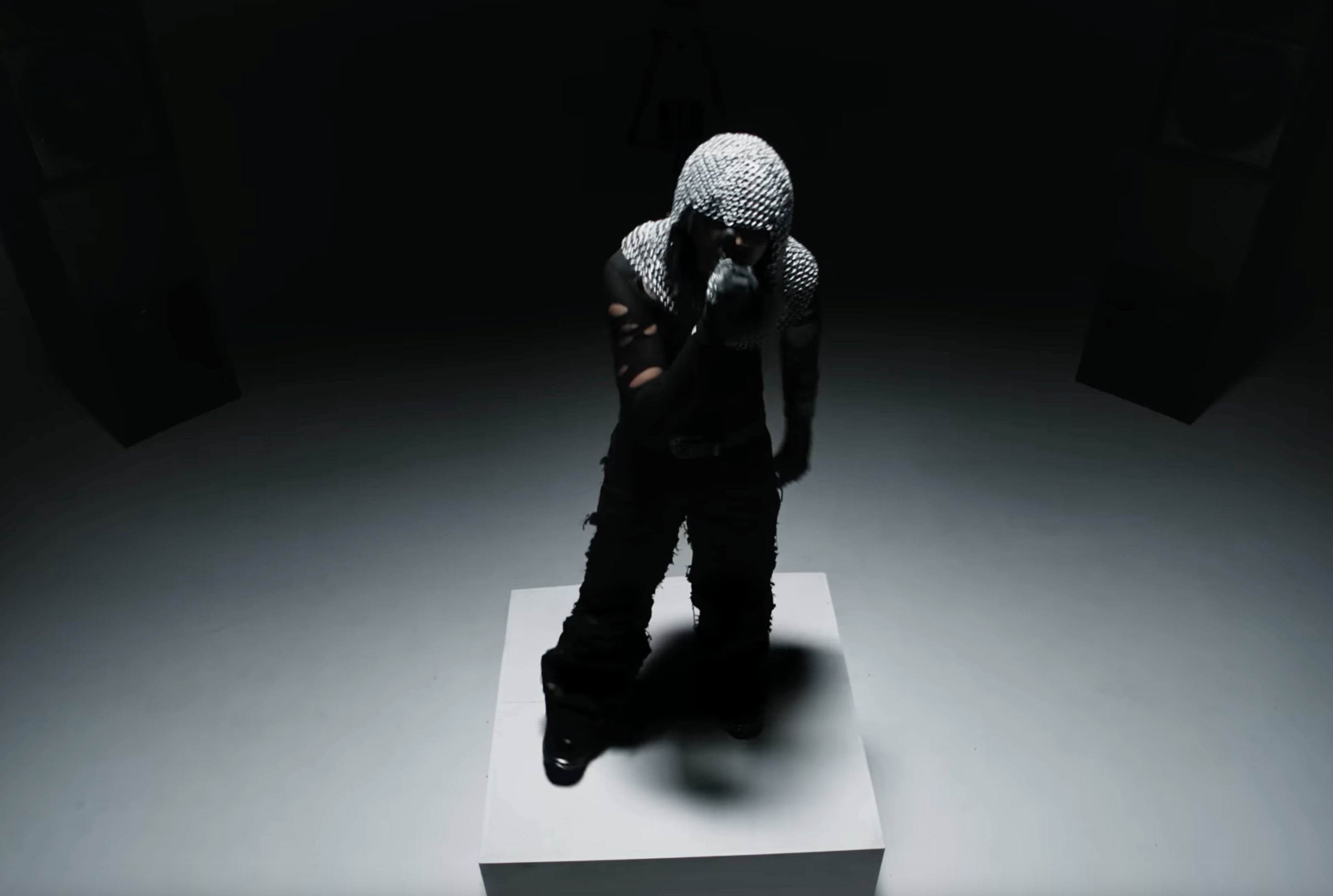 Ades The Planet wearing a chainmail hood and black torn clothing standing on a white square platform in a dimly lit room. - screenshot of the videoclip ADES THE PLANET, 24 Carats