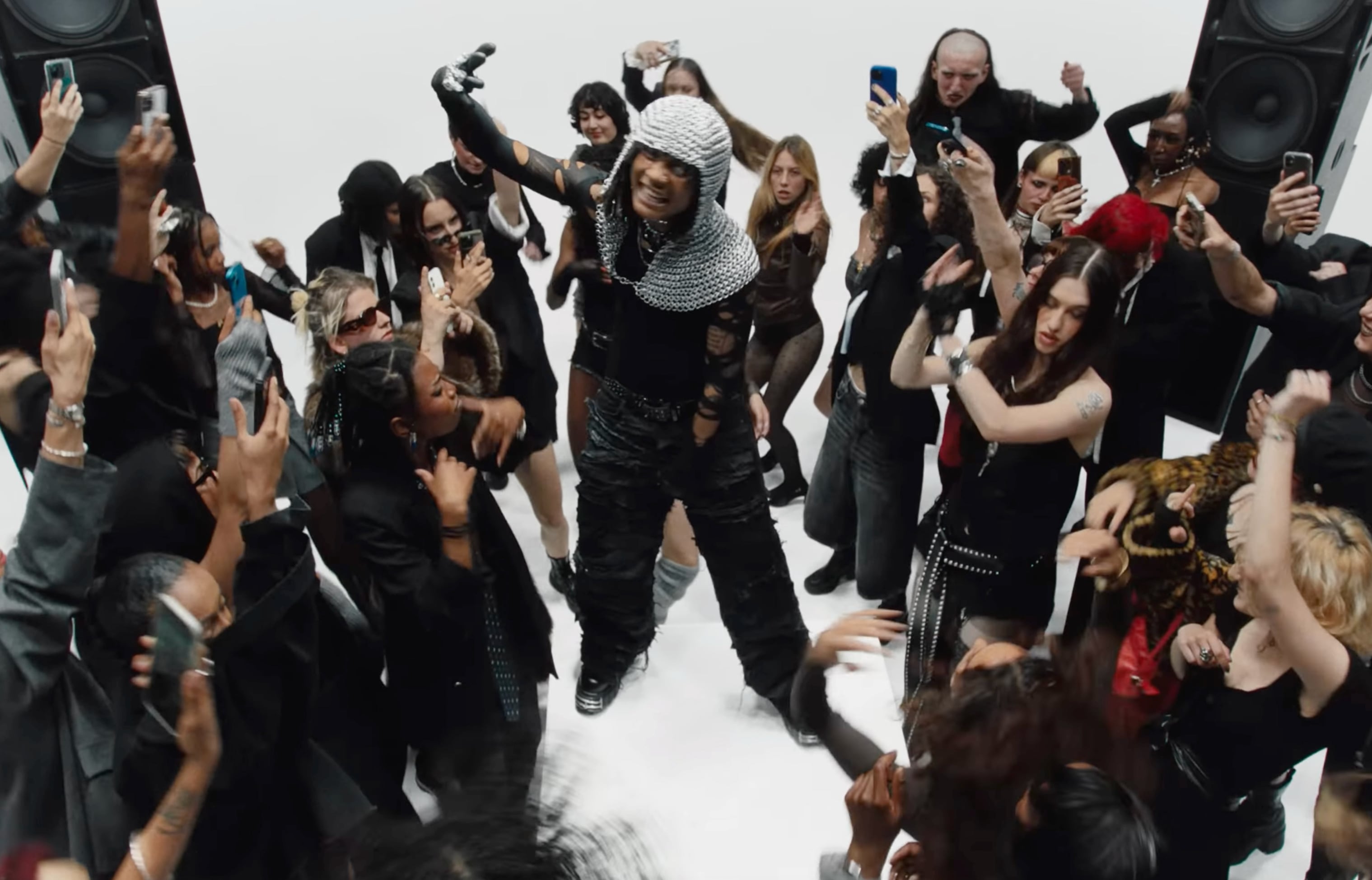 A diverse crowd dancing and taking photos around a performer wearing a chainmail hood and black outfit in a white studio setting. - screenshot of the videoclip ADES THE PLANET, 24 Carats
