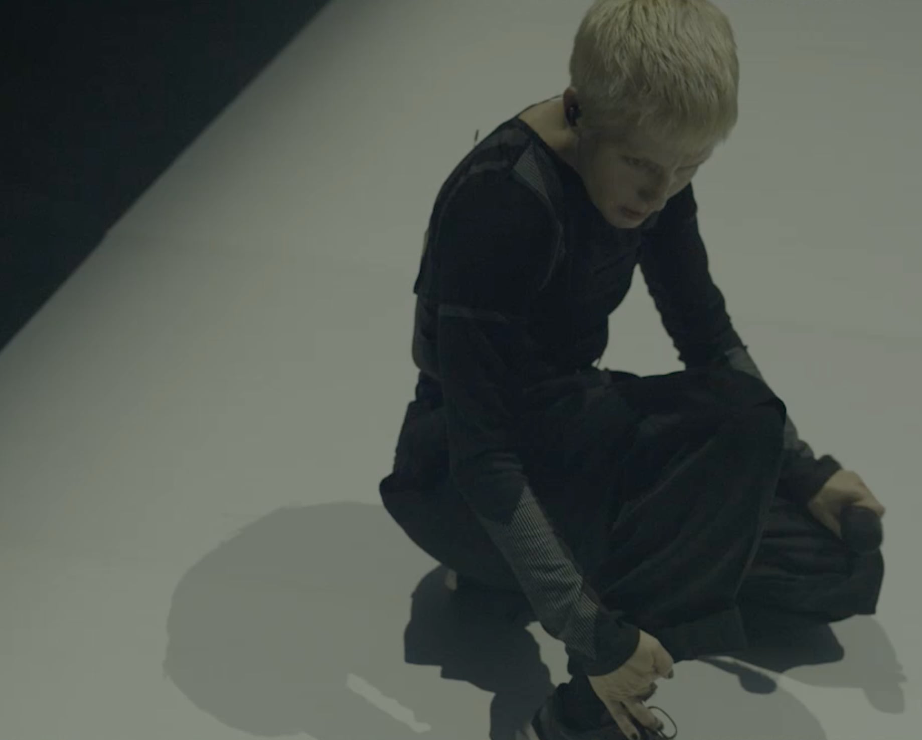 Jeanne Added with short blonde hair dressed in black crouching on a light-colored floor with a shadow cast beside them. - screenshot of the videoclip JEANNE ADDED, Both Sides