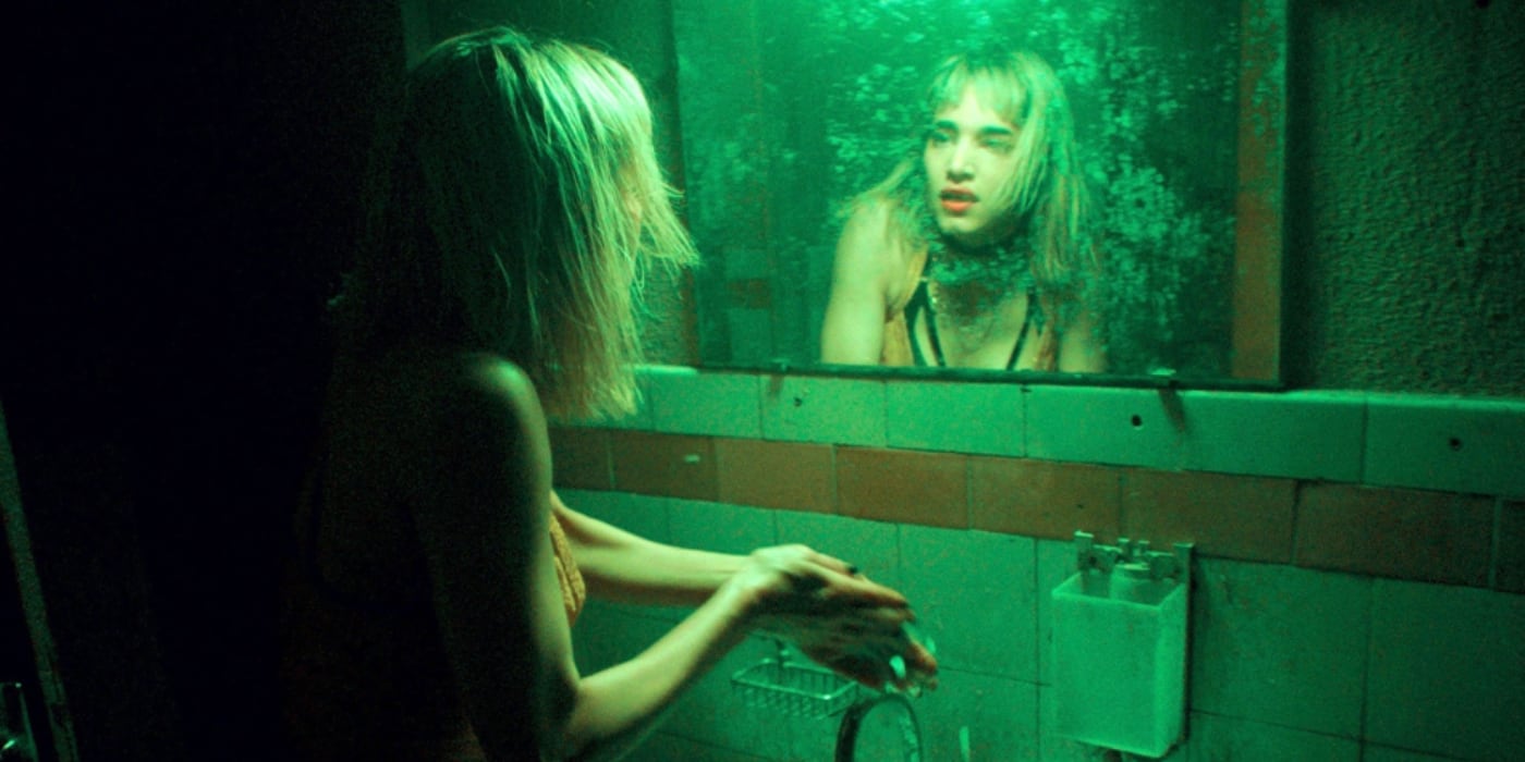 Person with wet hair washing hands in a dimly lit green-tinted bathroom, looking into a grimy wall mirror. - screenshot of the film CLIMAX