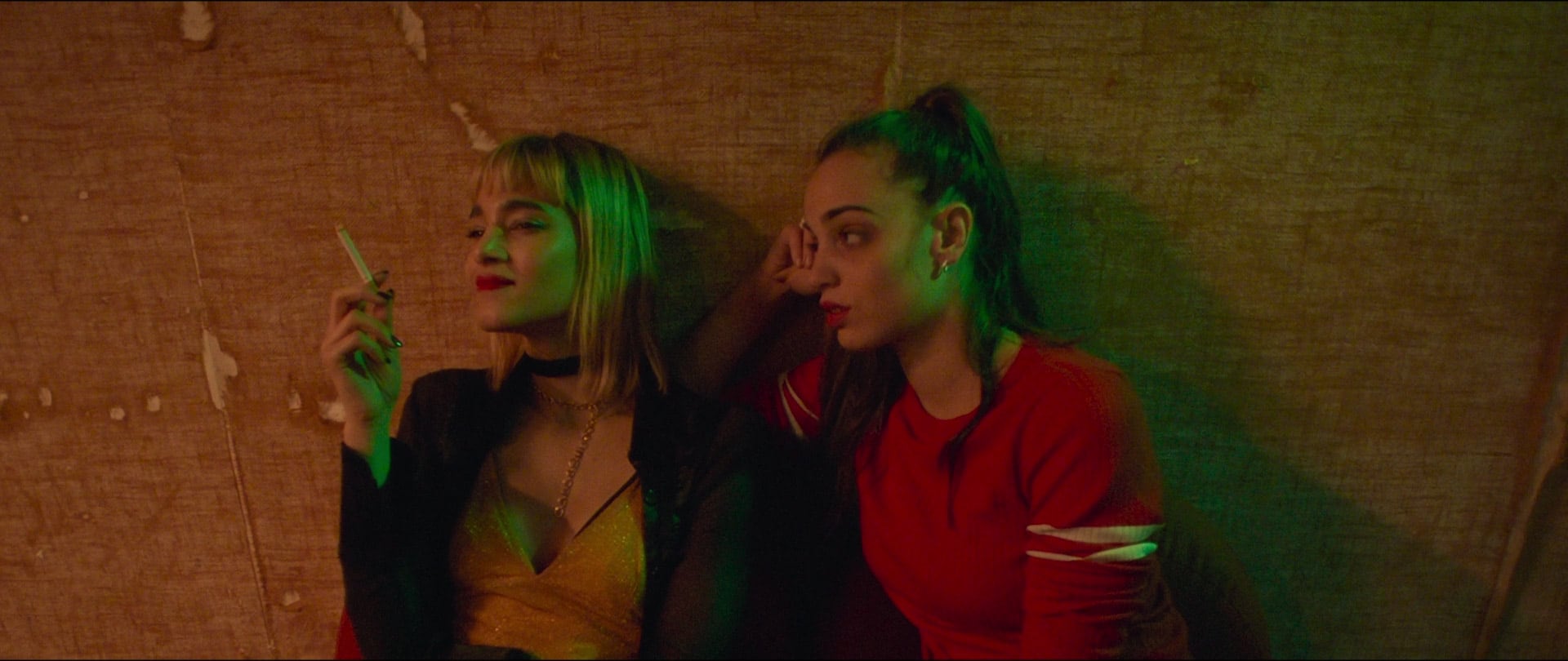 Two young women sit against a worn, brown wall with green lighting; the woman on the left holds a cigarette and wears a sparkly gold top and black jacket, while the woman on the right wears a red shirt looking to the left. - screenshot of the film CLIMAX