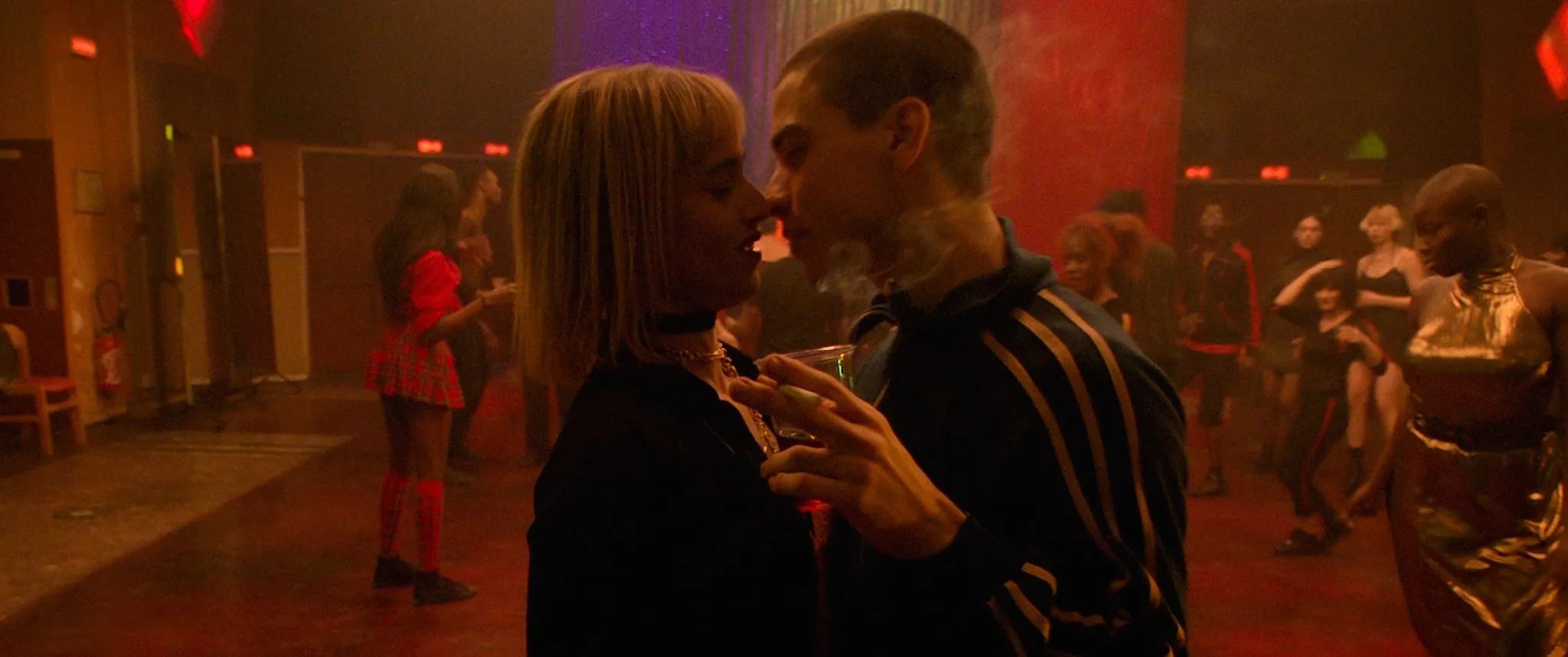 A couple stands close together in a dimly lit room with red and purple lighting, the man smoking a cigarette while holding a glass, surrounded by other people in the background. - screenshot of the film CLIMAX