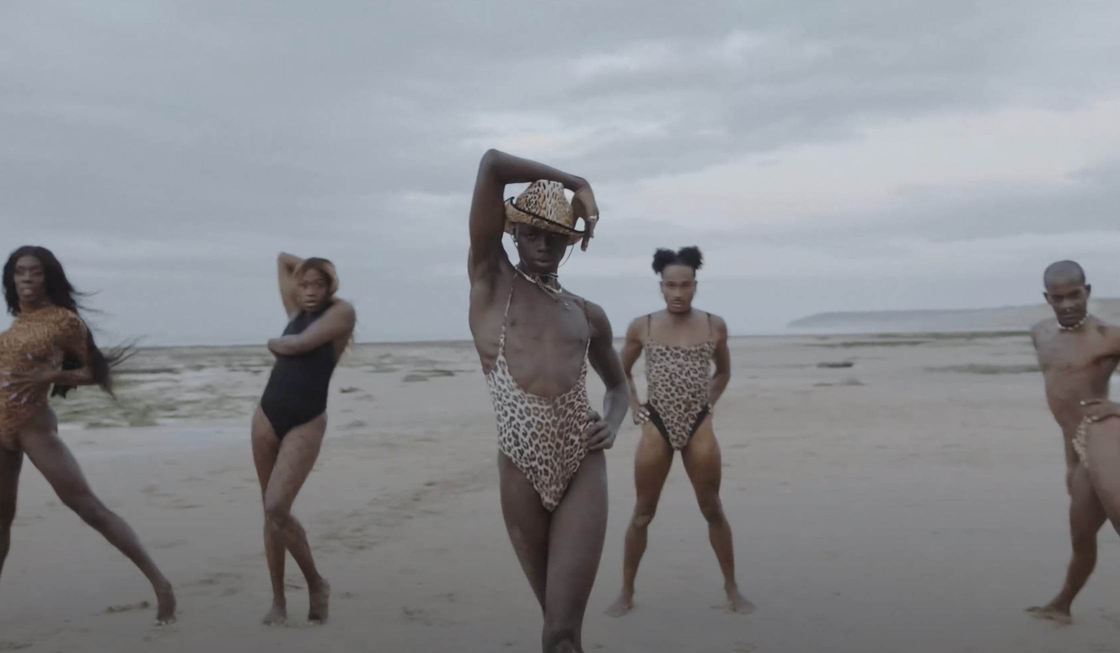 Five people posing confidently on a beach wearing leopard print and black swimsuits under a cloudy sky. - screenshot from the video NOWNESS, Tajabone - Just Dance