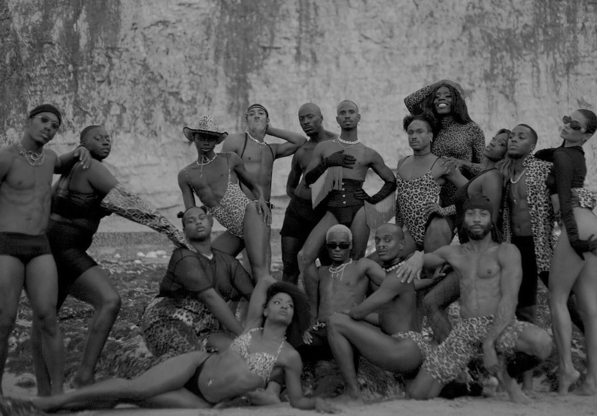 Group of fourteen people dressed in stylish leopard print and black outfits posing confidently on a rocky beach with a textured cliff background. - screenshot from the video NOWNESS, Tajabone - Just Dance