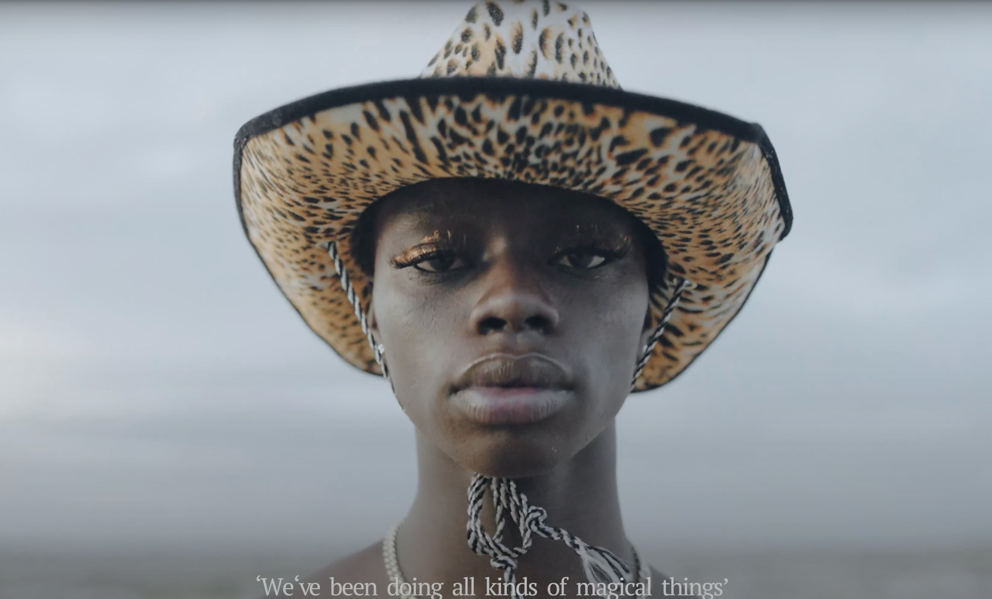 Close-up of a person with dark skin wearing a leopard-print cowboy hat and long eyelashes, against a muted gray sky. - screenshot from the video NOWNESS, Tajabone - Just Dance