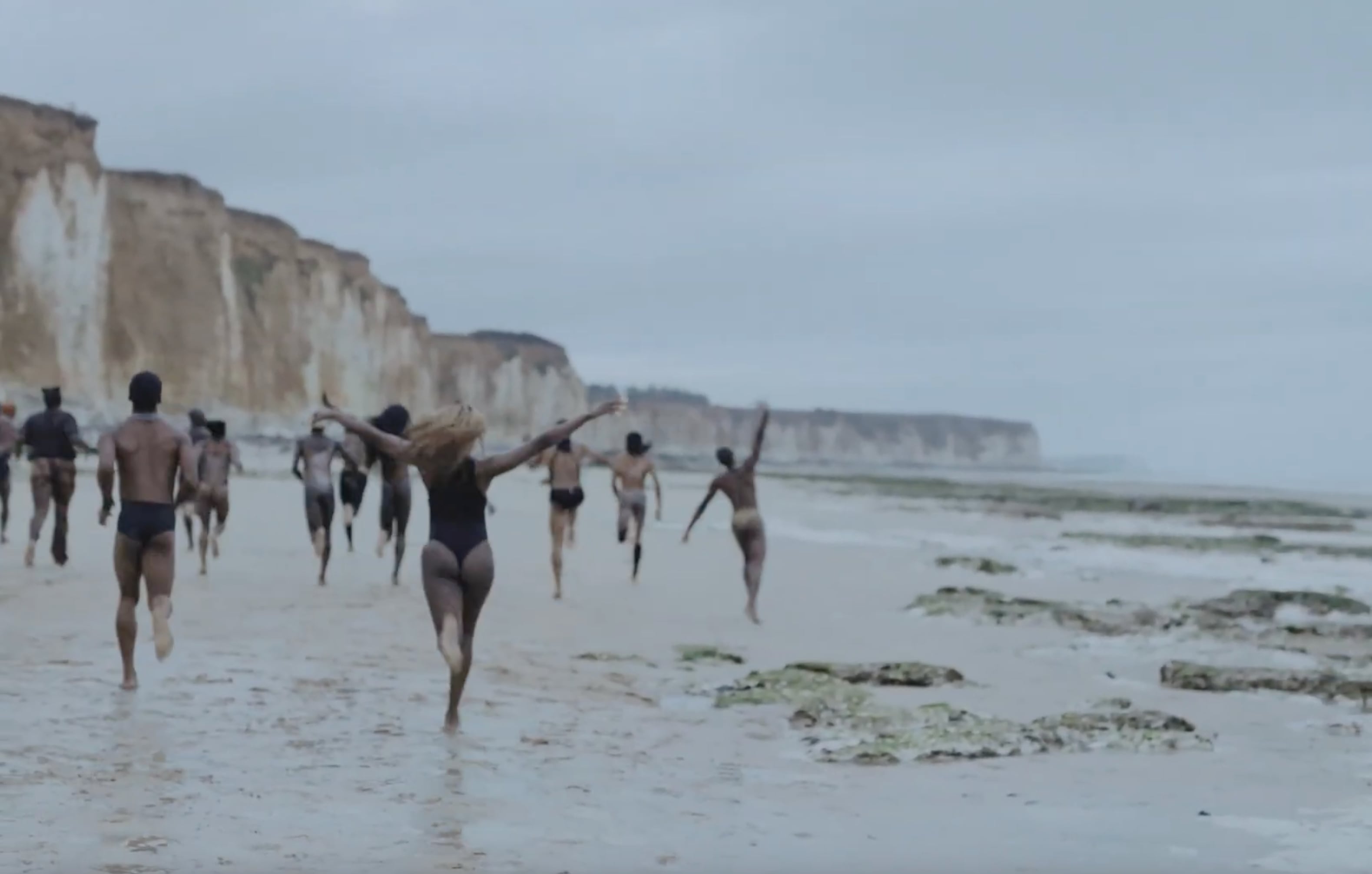 Group of people running and playing on a wet sandy beach with cliffs in the background under a cloudy sky. - screenshot from the video NOWNESS, Tajabone - Just Dance