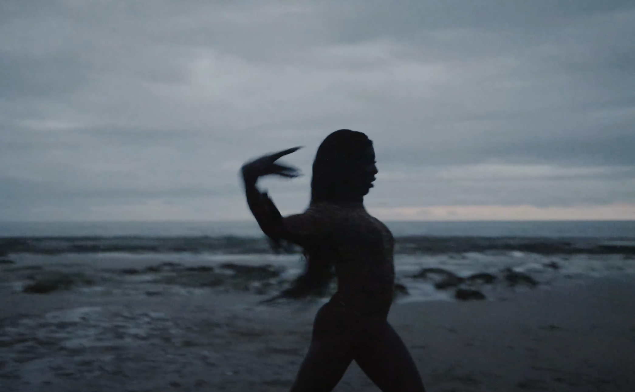 Silhouette of a person dancing on a rocky beach at dusk with waves and a cloudy sky in the background. - screenshot from the video NOWNESS, Tajabone - Just Dance