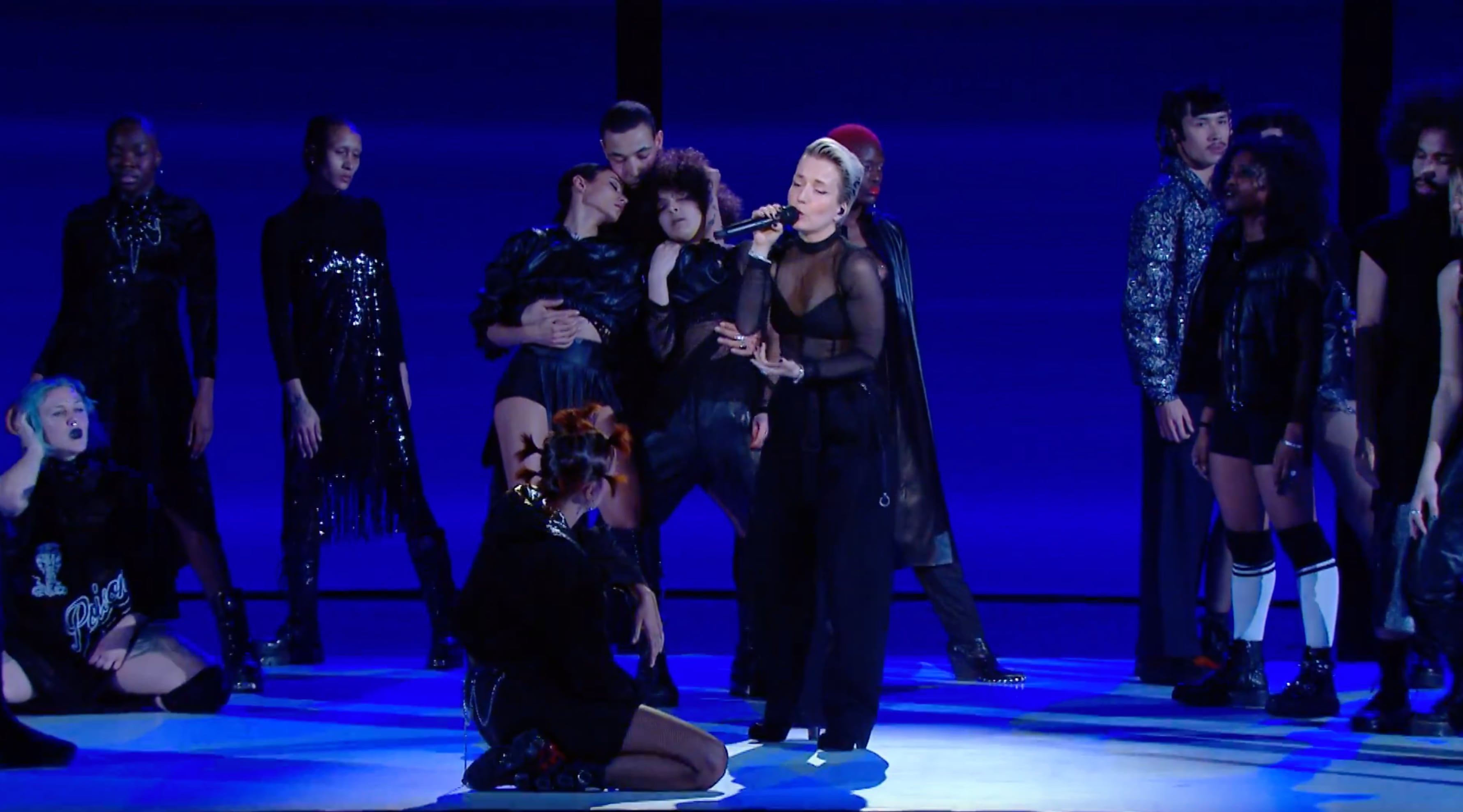 A diverse group of performers on stage with a purple-blue background, featuring a singer in a sheer black top holding a microphone surrounded by dancers in black outfits. - screenshot of the live performance of JEANNE ADDED x La Créole - Mutate during Les Victoires de la Musique