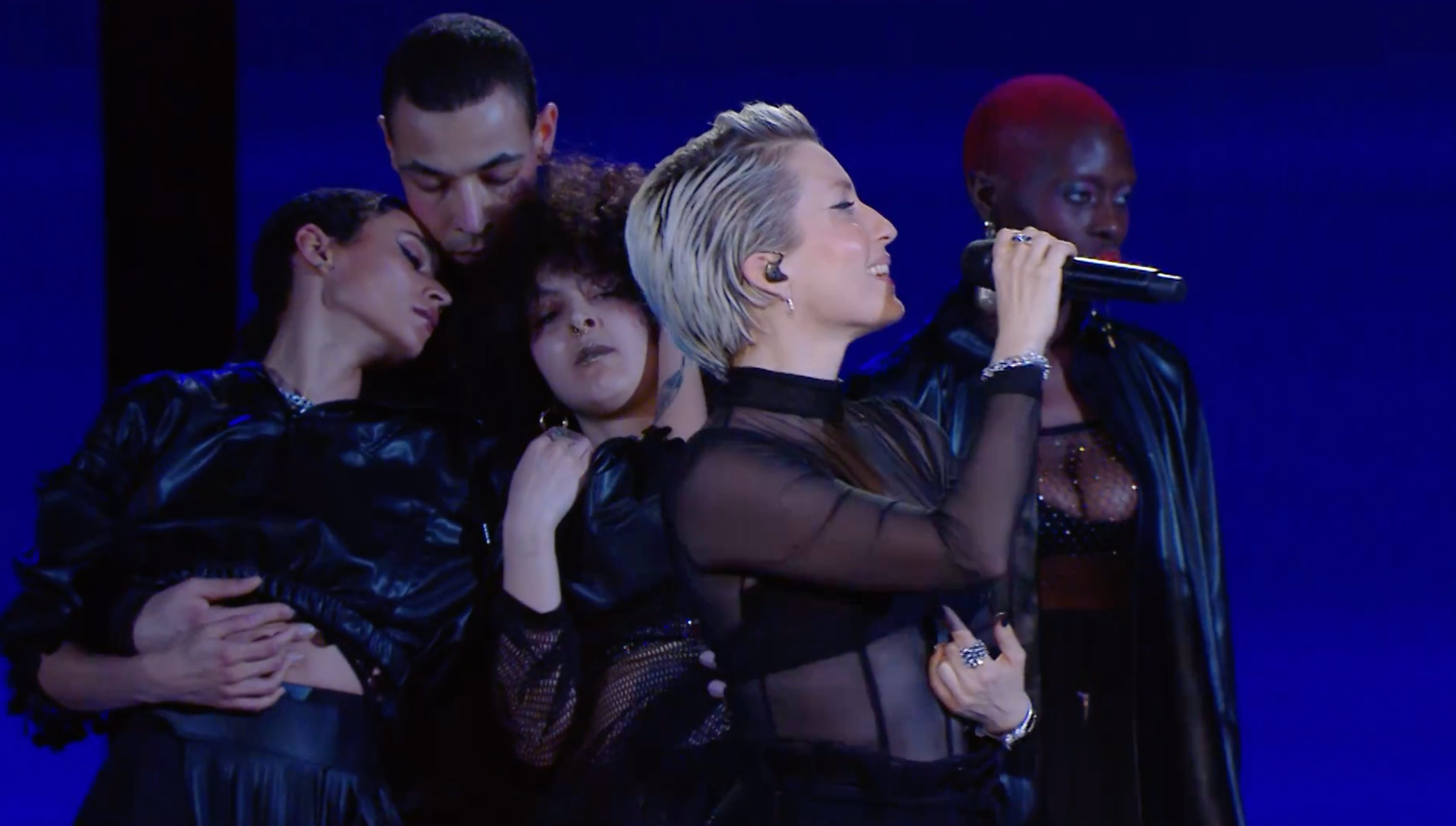 Five performers in black outfits on stage, with one singer holding a microphone and others closely grouped behind her against a dark blue background. - screenshot of the live performance of JEANNE ADDED x La Créole - Mutate during Les Victoires de la Musique