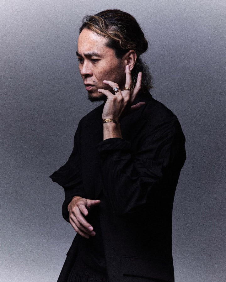 KOKI NAKANO with wavy hair tied back wearing a black jacket and multiple rings, touching his ear thoughtfully against a gray background.