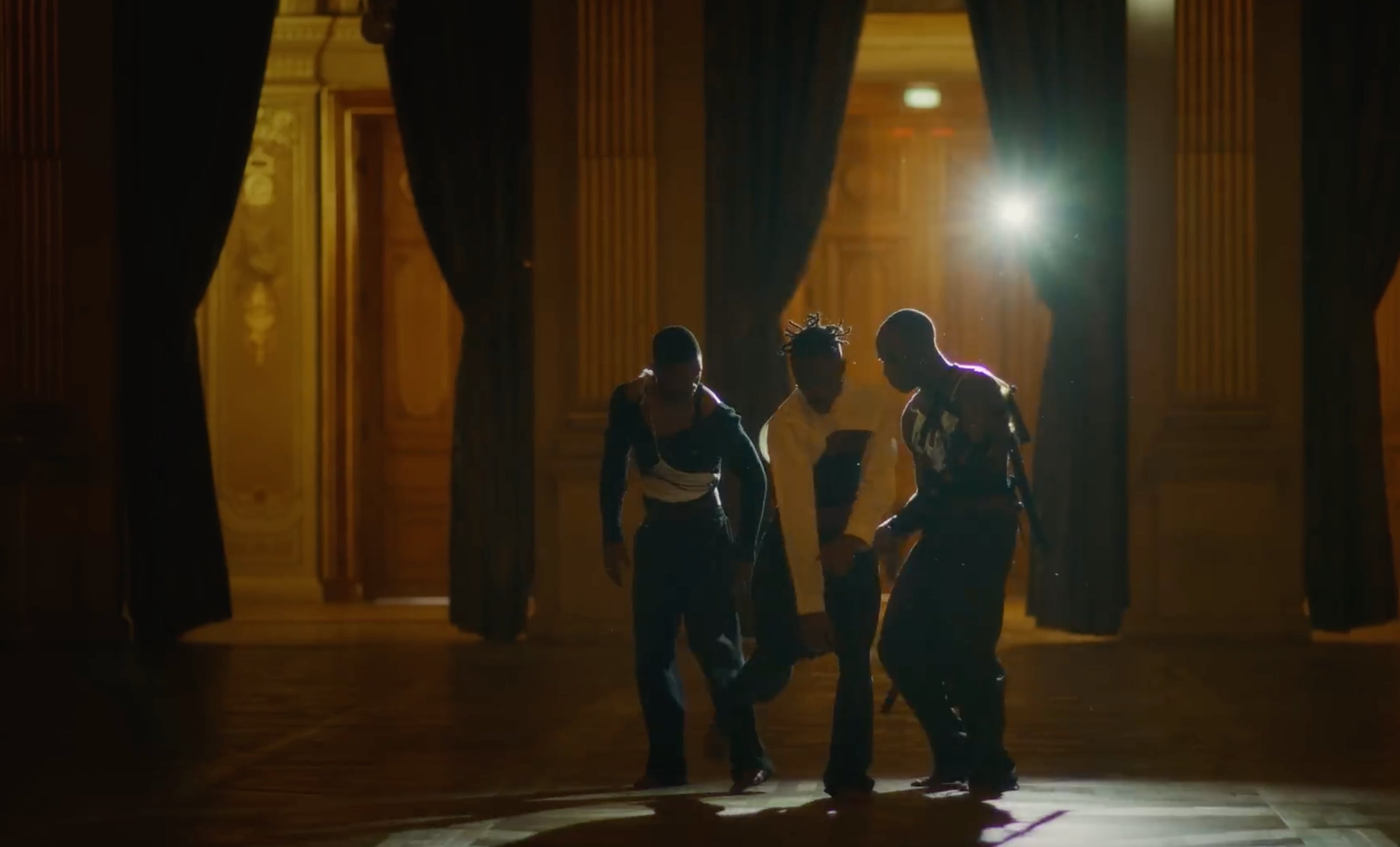 Three men dancing in a dimly lit ornate room with tall curtains and wood-paneled walls. - Screenshot of the video of KOKI NAKANO,
Mue