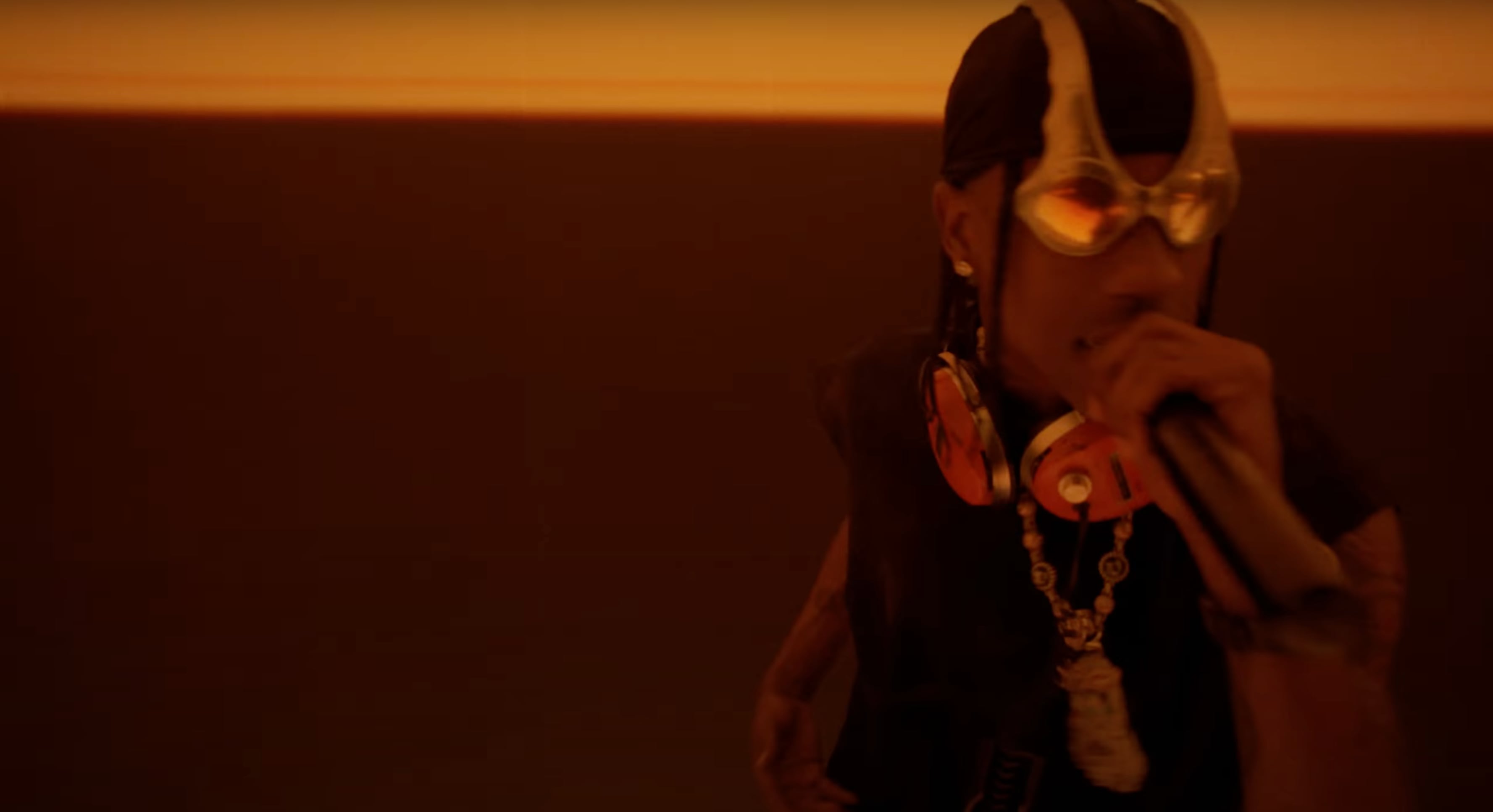 Person wearing reflective goggles and orange headphones around their neck, holding a microphone in a dimly lit orange environment. - Screenshot of the videoclip TRAVIS SCOTT, Modern Jam