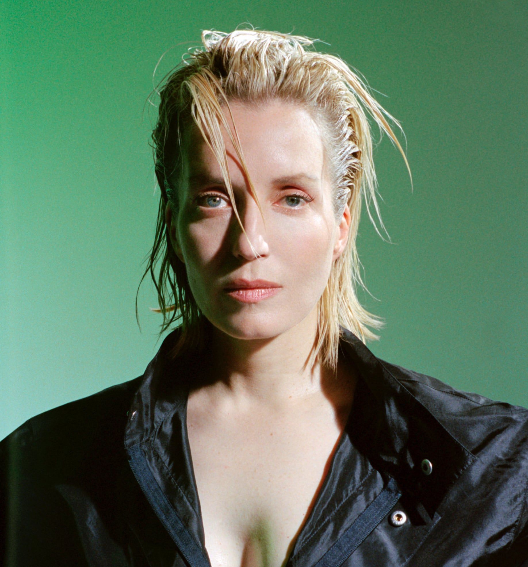 Close-up portrait of JEANNE ADDED with wet blonde hair, wearing a black jacket against a green background.