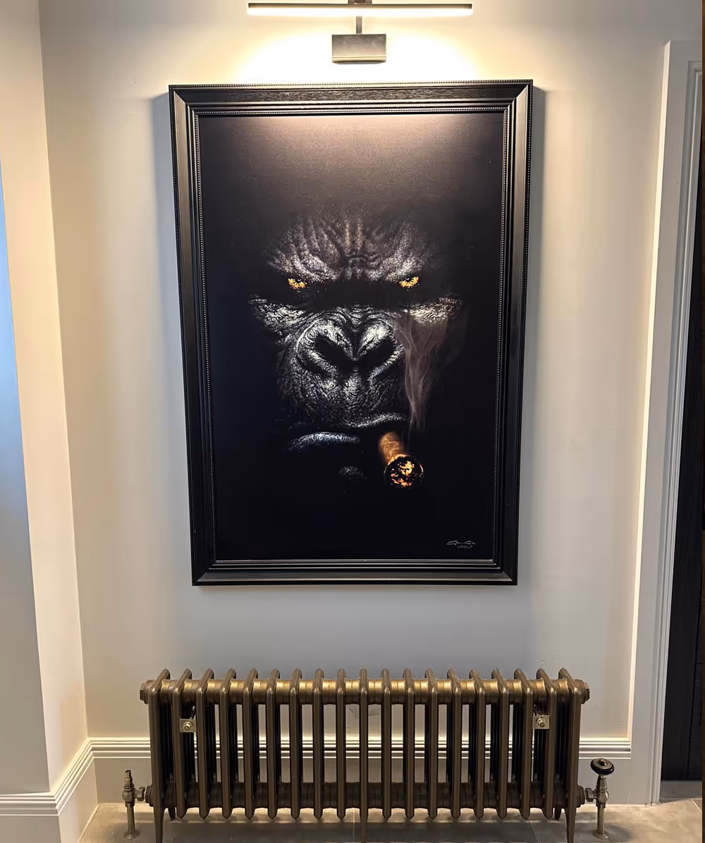 Framed painting of a gorilla with glowing yellow eyes smoking a cigar, mounted above a vintage radiator on a light-colored wall. Province Group Construction.