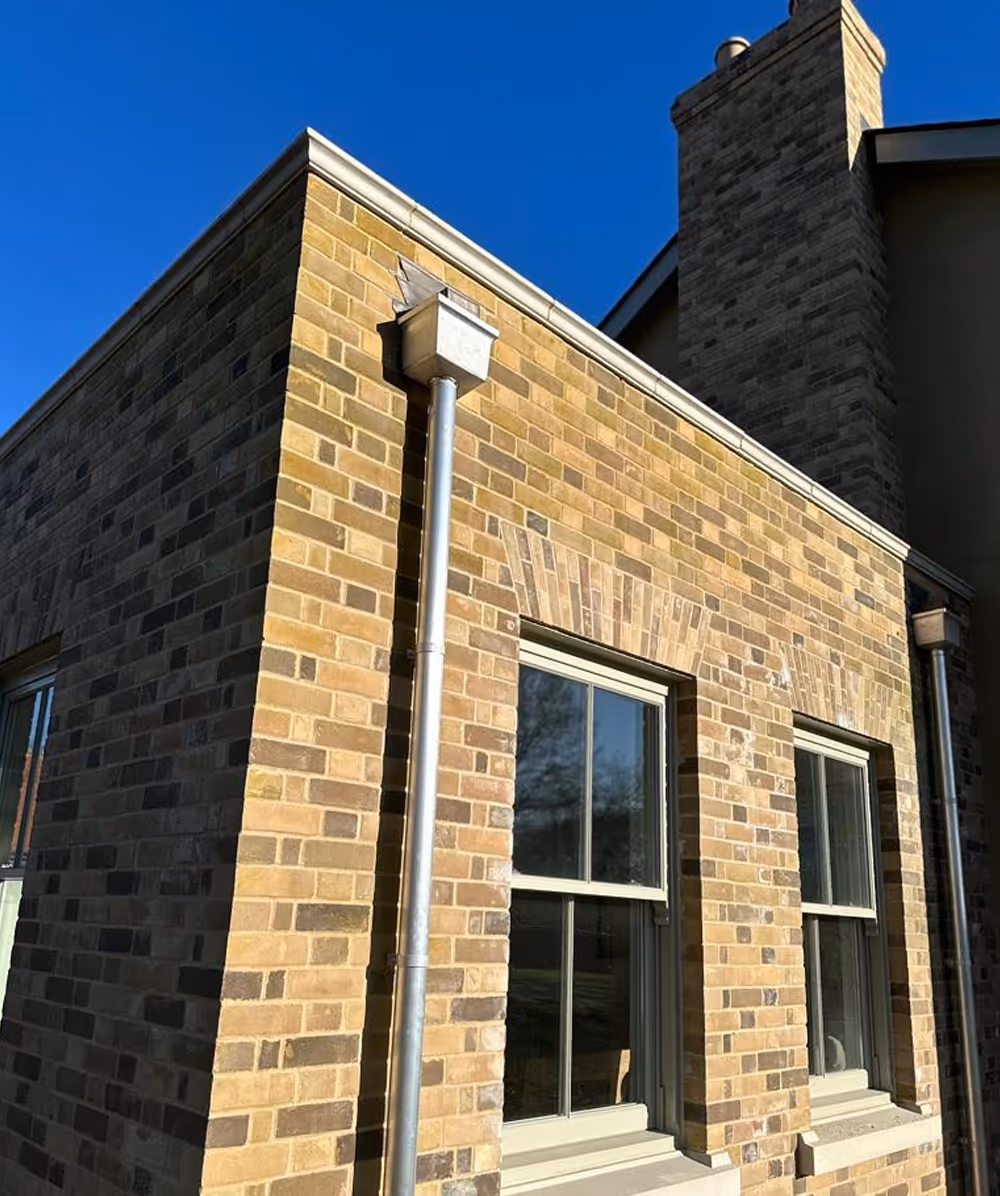 Corner of a brick building with two windows, metal downspouts, and a chimney under clear blue sky. Province Group Construction.
