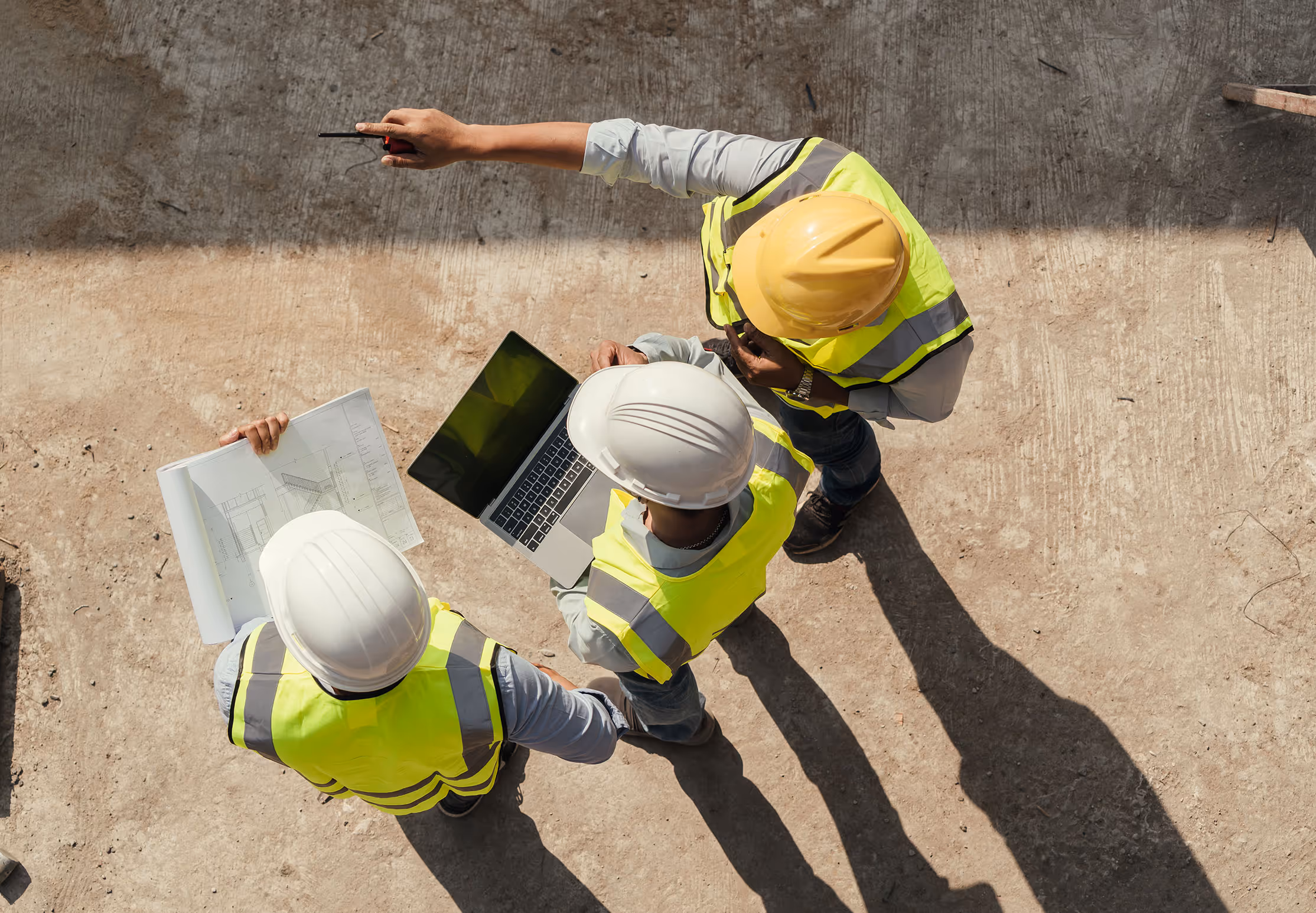 Three construction workers wearing white and yellow hard hats and reflective vests reviewing blueprints and a laptop on a concrete surface. Province Group Construction.