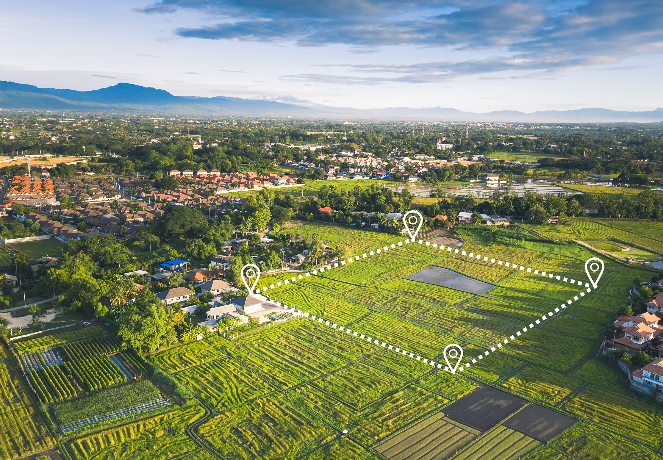 Aerial view of a green agricultural area with a marked rectangular plot outlined by white dotted lines and location pins, surrounded by houses and mountains in the distance. Province Group Construction.