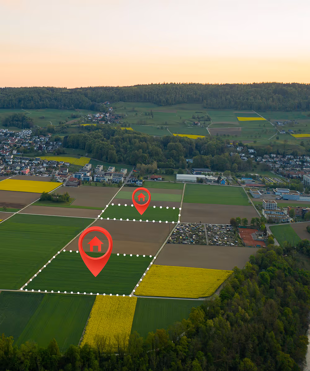 Aerial view of farmland with two marked plots outlined by white dotted lines and red location pins with house icons, near a town and forested area at sunset. Province Group Construction.