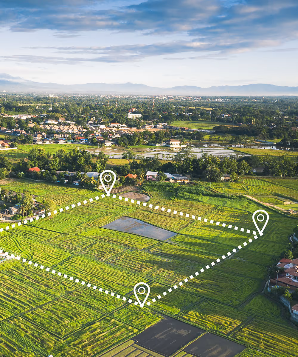 Aerial view of green agricultural fields with a white dashed boundary and three location markers, with a town and mountains in the background. Province Group Construction.