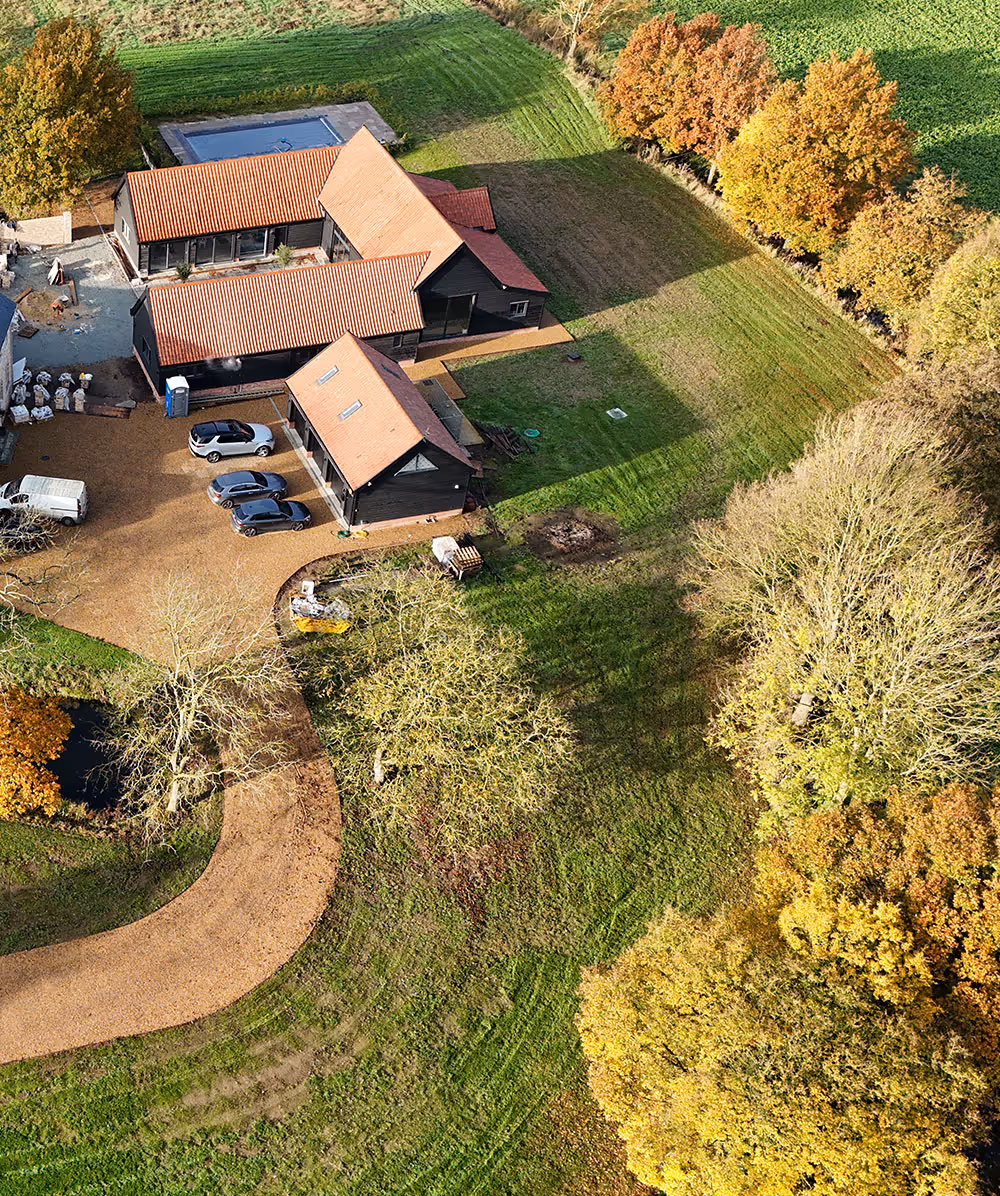 Aerial view of a farmhouse with red-tiled roofs, surrounded by green grass and trees with autumn foliage, and several parked cars near a curved gravel driveway. Province Group Construction.