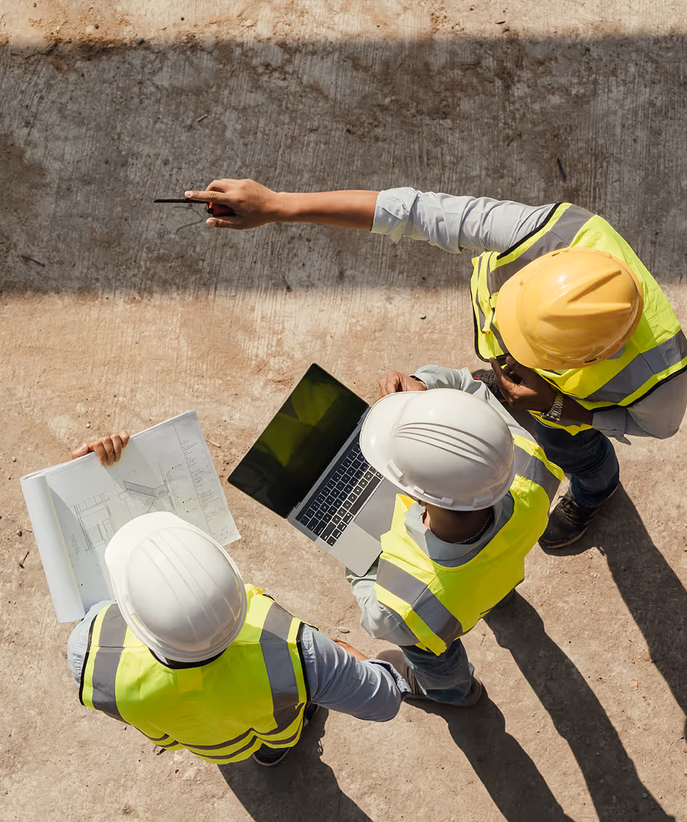 Three construction workers in safety vests and helmets reviewing a laptop and blueprints on a site. Province Group Construction.
