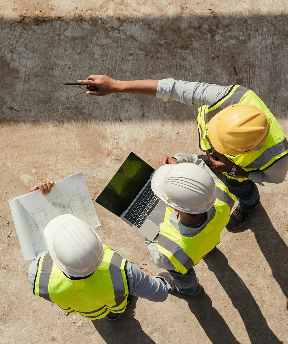 Three construction workers in safety vests and helmets reviewing a laptop and blueprints on a site. Province Group Construction.