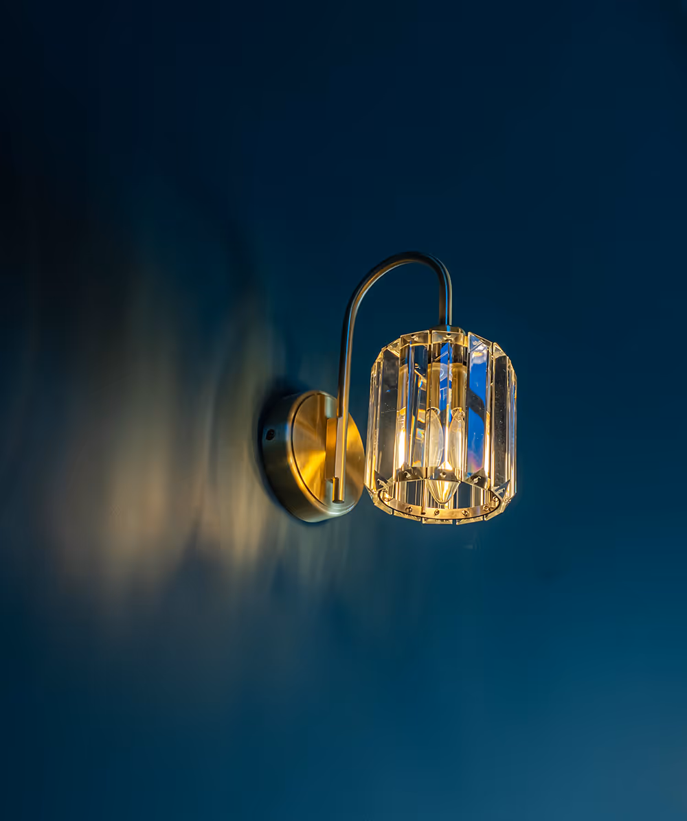 Wall-mounted brass light fixture with a faceted glass shade casting warm light on a dark blue wall. Province Group Construction.