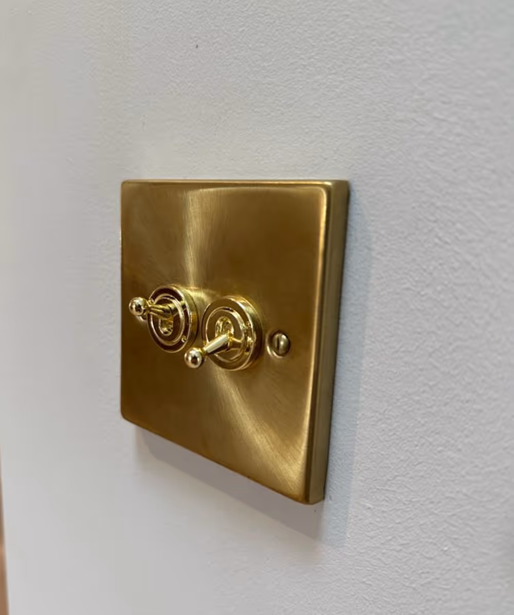 Brushed gold metal light switch plate with two toggle switches on a white textured wall. Province Group Construction.
