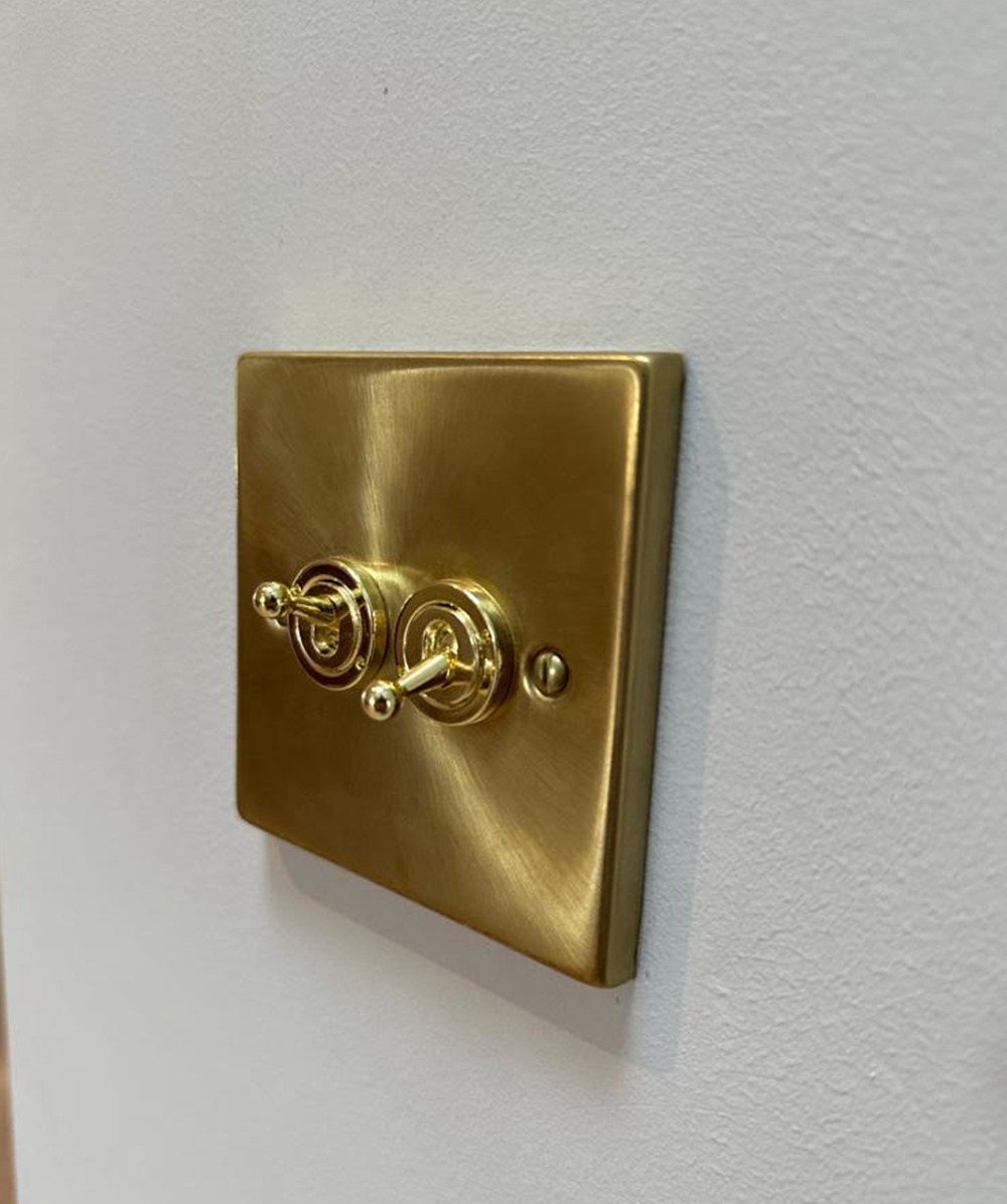 Brushed gold metal light switch plate with two toggle switches on a white textured wall. Province Group Construction.