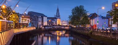 Evening cityscape with a river, lit buildings, and a church with a tall spire in the background.