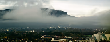 Mountain partially covered by fog over a town with buildings and trees in the foreground.