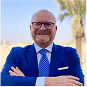 Smiling bald man with glasses in blue suit and tie, arms crossed outside on a sunny day.