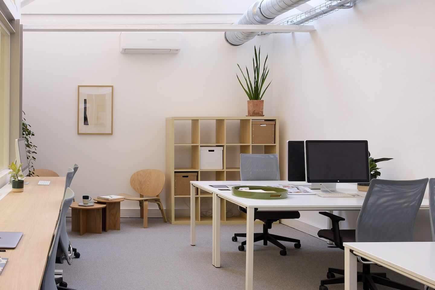 Bureau moderne et minimaliste avec chaises de bureau grises, deux écrans d’ordinateur, étagère beige avec plantes et boîtes de rangement, murs blancs et tapis gris.