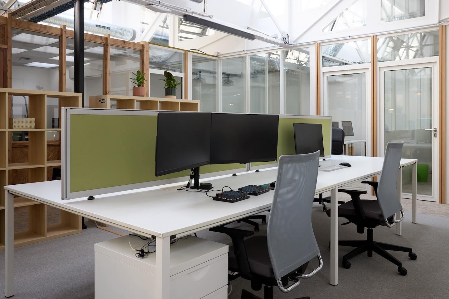 Bureau moderne ouvert avec deux postes de travail équipés de plusieurs écrans, chaises de bureau grises et séparateur vert clair.