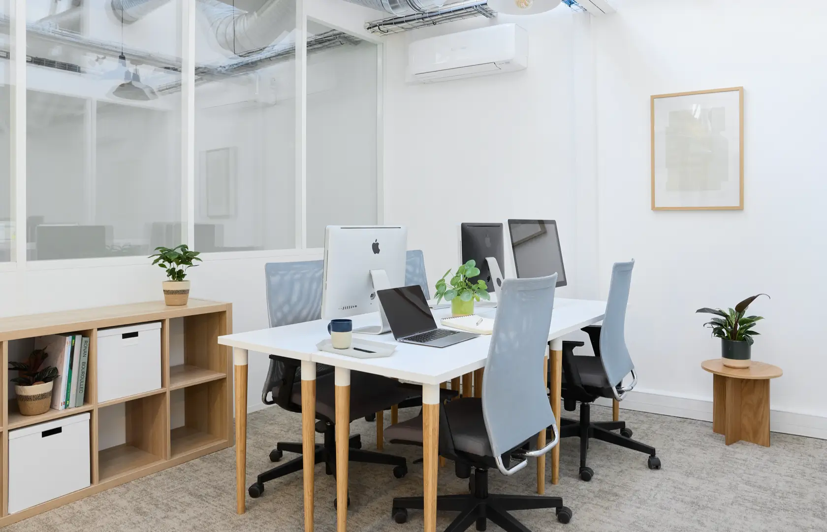 Bureau moderne avec quatre ordinateurs, chaises grises, plantes décoratives, et étagère en bois avec rangement.