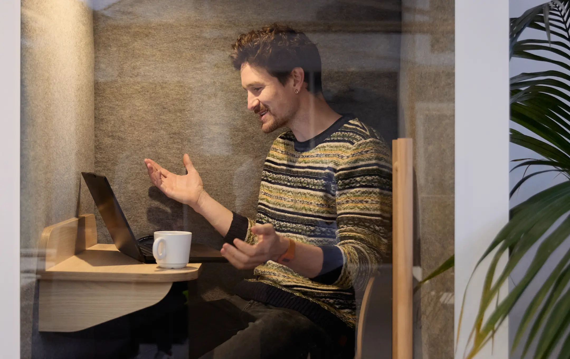 Un homme souriant discutant en ligne dans une cabine de bureau isolée avec un ordinateur portable et une tasse blanche.